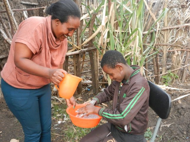Woman pouring water into bucket, as young boy washes hands in the bucket.