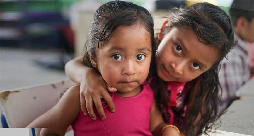 A young girl with her arm around another young girl with a disability.