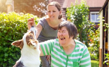 A smiling female with a disability feeding a white and brown-haired dog a treat while another female stands behind her.