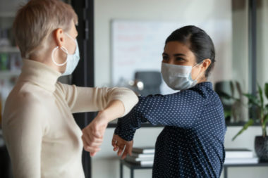 Two female colleagues wearing blue non medical masks while bumping elbows in the workplace.
