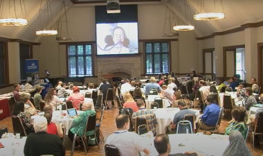 Several groups of people sitting at tables in a conference centre watching a video at the front of the room.
