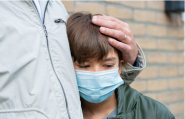 Young boy with a blue non medical mask standing closely to his father while his father holds his head.