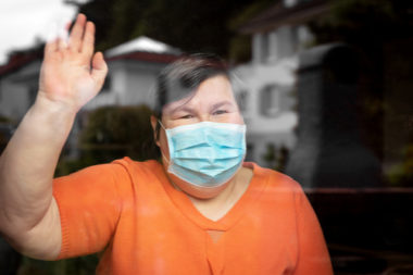 Woman with disability wearing a blue non medical mask while waving at someone through a window.