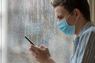 Man wearing a blue non medical mask while looking at his cellphone in front of a window.