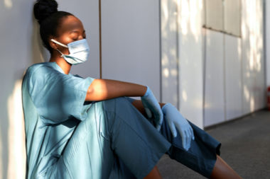 Nurse sitting outside wearing scrubs, a blue non medical mask, and latex gloves.